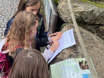 Drei Schülerinnen an einer Steinwand beim Ausfüllen der Fragenrallye